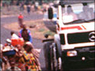 Three military vehicles pass alongside walking villagers on a narrow road.