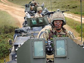A group of men, each wearing military gear, drive armoured vehicles down a dirt road.