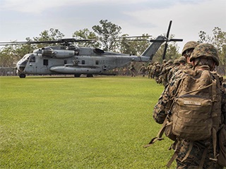 A photograph of a line of Marines wearing camouflage walking across a grass field to board a large helicopter.