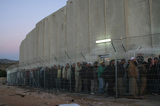 Daybreak, line of people behind a fence adjacent to cement wall.