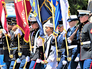 A group of six men and two women, all-uniformed, with some carrying colorful flags, marches in formation.