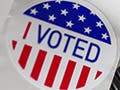 A strip of red, white, and blue stickers with the words “I Voted” lies next to an announcement board with the words “Voting Day” written upon it.