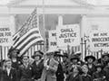 Wives of political prisoners standing in a crowd with protest signs and an American flag.