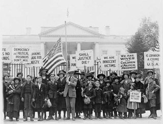 Wives of political prisoners standing in a crowd with protest signs and an American flag.