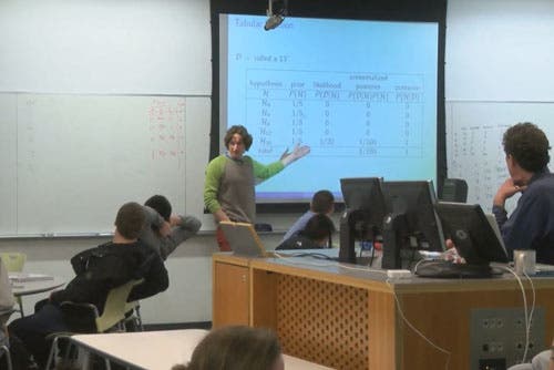 An instructor points at a projector screen showing a slide and addresses students, who are sitting at round tables.