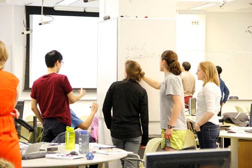Small groups of students gather around whiteboards and work together on solving math problems.