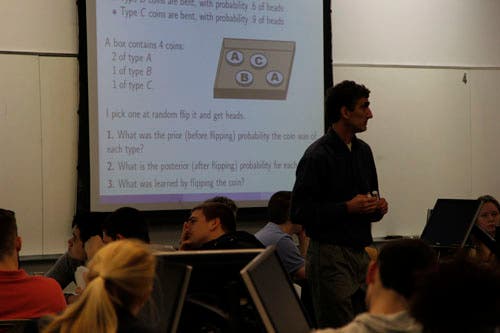 An instructor stands amidst seated students. The screen on the wall shows a slide with a math problem for the students to work on.