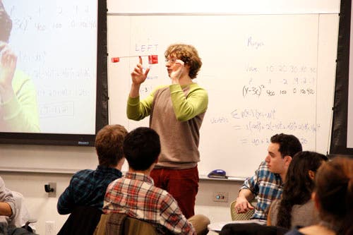 An instructor balances a ruler with weights on his finger, as students look on.