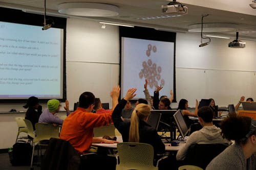 Students seated at round tables raise their hands. One projector screen shows a slide and one projector screen shows a set of dice.