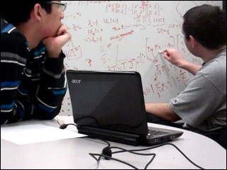 Two students working on a number of mathematical equations on a whiteboard.