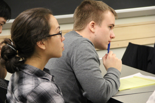 Three students sitting at a table; female student looks toward speaker at front of room; male student leans forward, holds pen, and looks toward speaker; another male student looks down at paper.