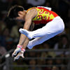 A male gymnast is in a straddled position midair with his hands reaching down to catch the high bar.