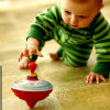 An infant on hands and knees reaches out an exploratory hand toward a large toy spinning top in motion on the floor in front of it.