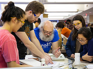 Four students and one professor leaning over a table pointing and looking at items and papers on the table.