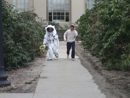 One astronaut and one crew member practice loping along the sidewalk while communicating via walkie-talkie.