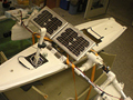 A long slim white hull sits in a cradle, with PVC/Styrofoam pontoons extending to the sides, and a solar panel and periscope assembly sitting on top.