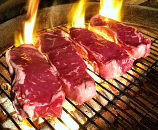 Four steaks on a charcoal grill with flames.