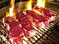 Four steaks on a charcoal grill with flames.