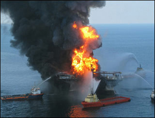 A large offshore drilling rig is up in flames. Several small ships are spraying it with water.