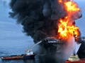 A large offshore drilling rig is up in flames. Several small ships are spraying it with water.