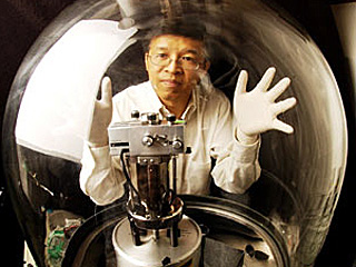 A man wearing white latex gloves posing both hands on a glass chamber containing a mechanical device.