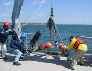 Three people winching large tripod off deck into ocean.