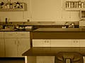 Photo of a biology lab bench, with two chairs, and shelves of chemical bottles in the background.