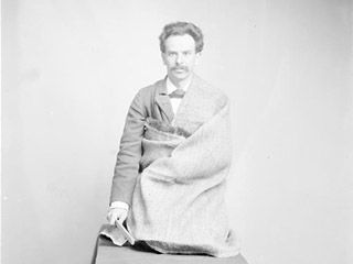 A young man with wavy hair and a bushy moustache, with burlap draped around his body, kneels on a table.