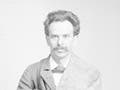 A young man with wavy hair and a bushy moustache, with burlap draped around his body, kneels on a table.