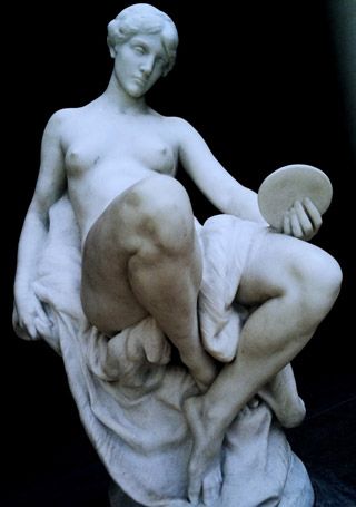 A marble statue of naked woman seated on a pedestal, legs crossed at the ankles, with linen draped between her thighs.
