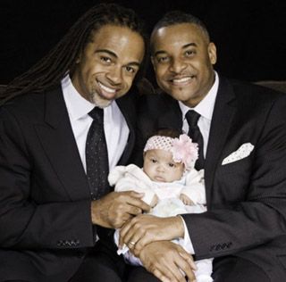 Two handsome, well-dressed men of color hold an infant, with a pink bow around her head.