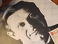 On a wooden tabletop, a black and white photograph of a distinguished-looking man wearing a bow tie rests beneath a pile of reel to reel tape boxes.