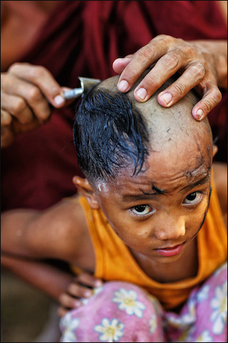 A photograph of a young Burmese boy who is in the middle of having his head shaved by an older monk.