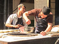One man stands near a huge pot while three other men prepare food for cooking.