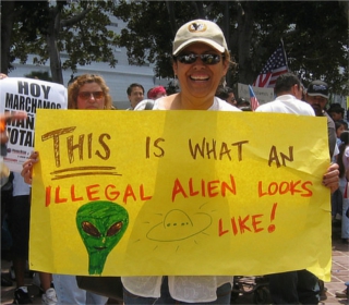Photo of a woman in a baseball cap holding a bright yellow sign reading "THIS is what an illegal alien looks like!", with a sketch of a green bug-eyed alien next to a flying saucer.