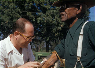 A white doctor gives a shot in the arm of an elderly black man.