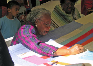 An elderly brown-skinned woman with gray hair and wearing glasses works on a quilt.