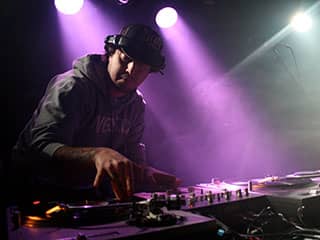 A young man wearing headphones stands behind two turntables.