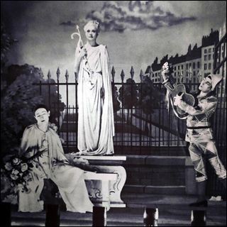 Film still: Onstage in a theater, a live tableau including a mime, a harlequin playing guitar, and Aphrodite in a public garden.