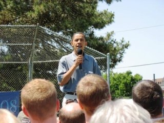 Barack Obama holding a microphone and speaking outdoors in front of a number of people.