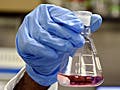 A young male, wearing goggles and gloves, observes  purple liquid in a beaker.