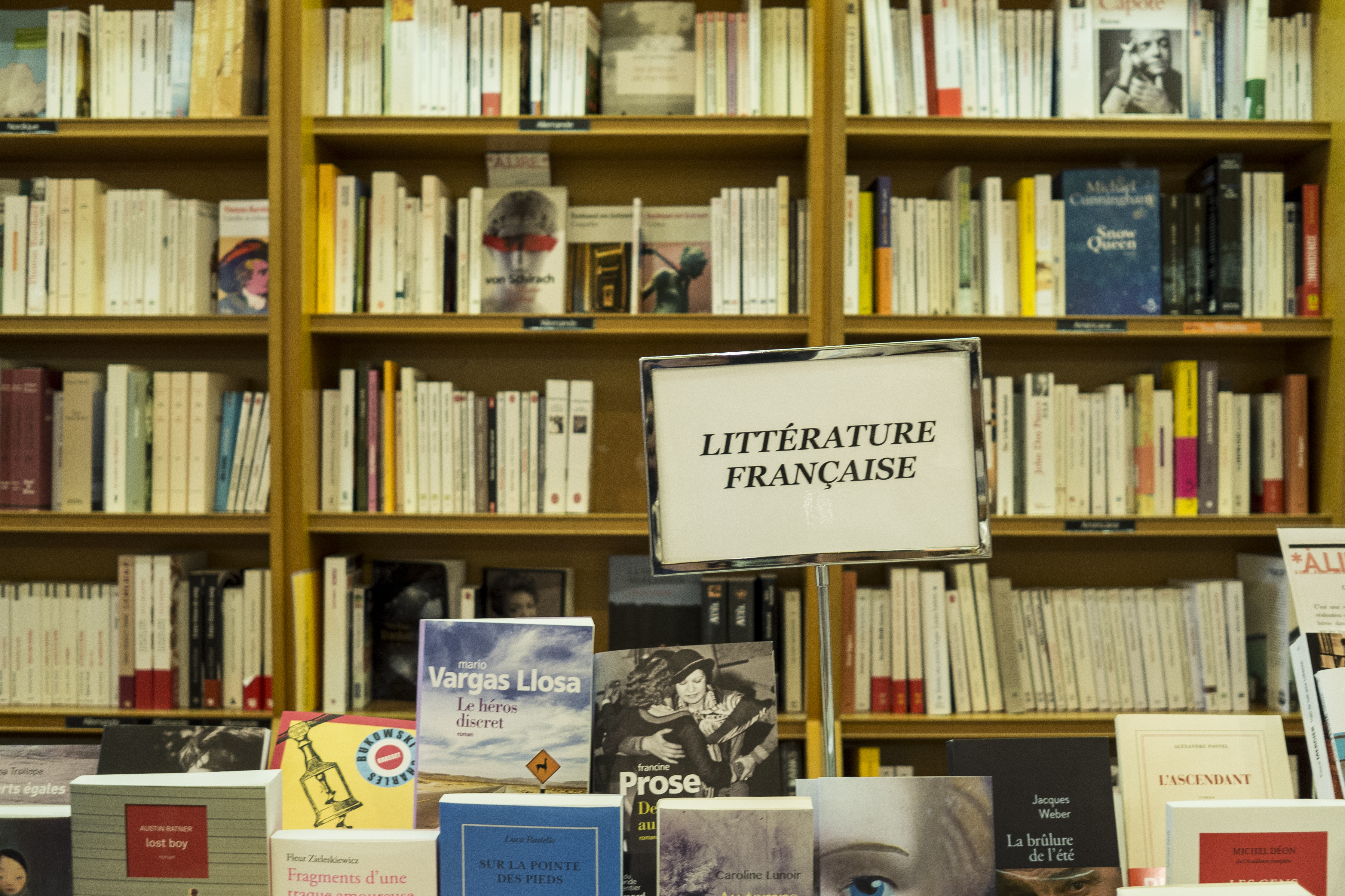 A view of several bookshelves in a bookstore stocked and organized neatly behind a row of display books with covers facing forward, a slightly askew metal sign-holder reads "littérature française."