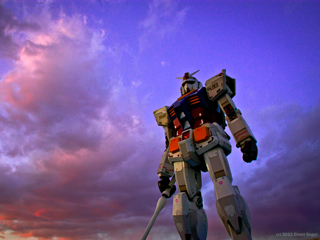A giant robot standing tall holding a saber in his hand with a purple sunset sky and pink clouds as the background.