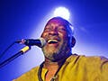 A man in a colorful green and yellow shirt performs on stage. He sits in front of a microphone with arms spread wide and has a hug grin on his face. 
