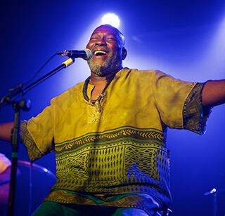 A man in a colorful green and yellow shirt performs on stage. He sits in front of a microphone with arms spread wide and has a hug grin on his face. 