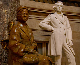 A bronze statue of a seated woman with her hands folded close to her purse is positioned near the marble statue of a standing gentleman wearing a knee-length coat, with one hand on his hip. 