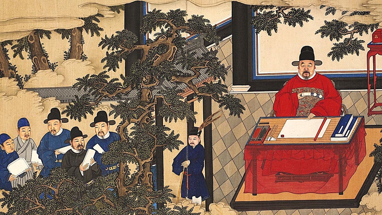 A man, with a thin goatee and wearing an elaborate red robe, sits at a desk while a group of men observe.