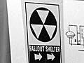 An attractive woman stands in front of a display featuring individually-sized supplies for a fallout shelter, such as food, and medical and sanitation kits.
