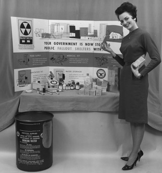An attractive woman stands in front of a display featuring individually-sized supplies for a fallout shelter, such as food, and medical and sanitation kits.