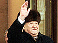 A grey-haired gentleman wearing a fur hat and black overcoat, raises his hand in a wave.